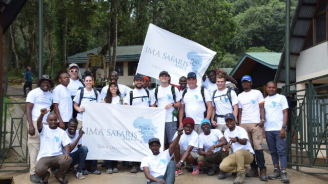 A group of people in matching shirts pose with flags labeled "IMA Safaris," possibly related to east Africa safari tours.