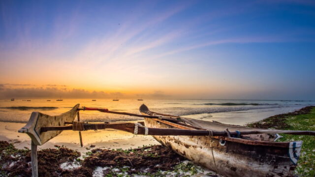 A traditional wooden boat on a sandy beach at sunrise, with a vibrant sky. Perfect for highlighting east africa safari tours.