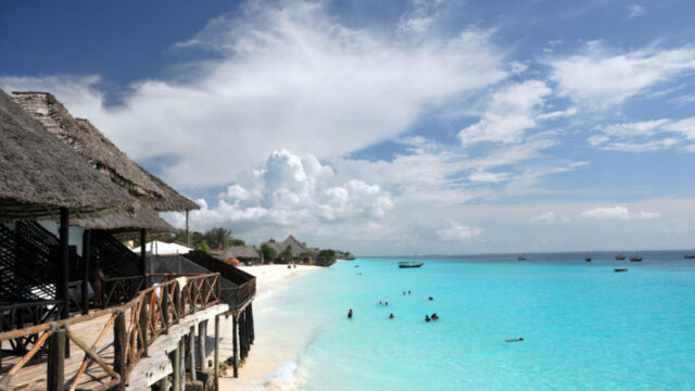 A tropical beach scene with clear turquoise water, people swimming, and thatched-roof structures along the shore.