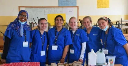 Group of six smiling healthcare interns in blue scrubs standing together in a classroom setting with medical supplies on the table in front of them.