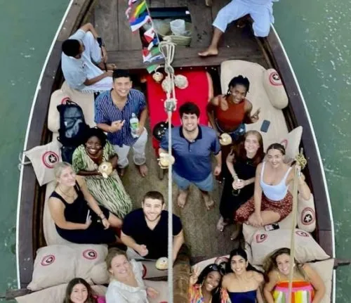 Group of smiling young adults relaxing on a decorated boat ride, enjoying drinks and scenic views during an outing on the water.