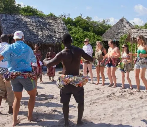 Group of travelers participating in a traditional dance lesson on a sandy beach, led by local performers in a coastal village setting.