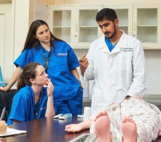 Medical instructor in a white coat teaching a group of attentive interns in blue scrubs using a mannequin during a clinical skills training session.
