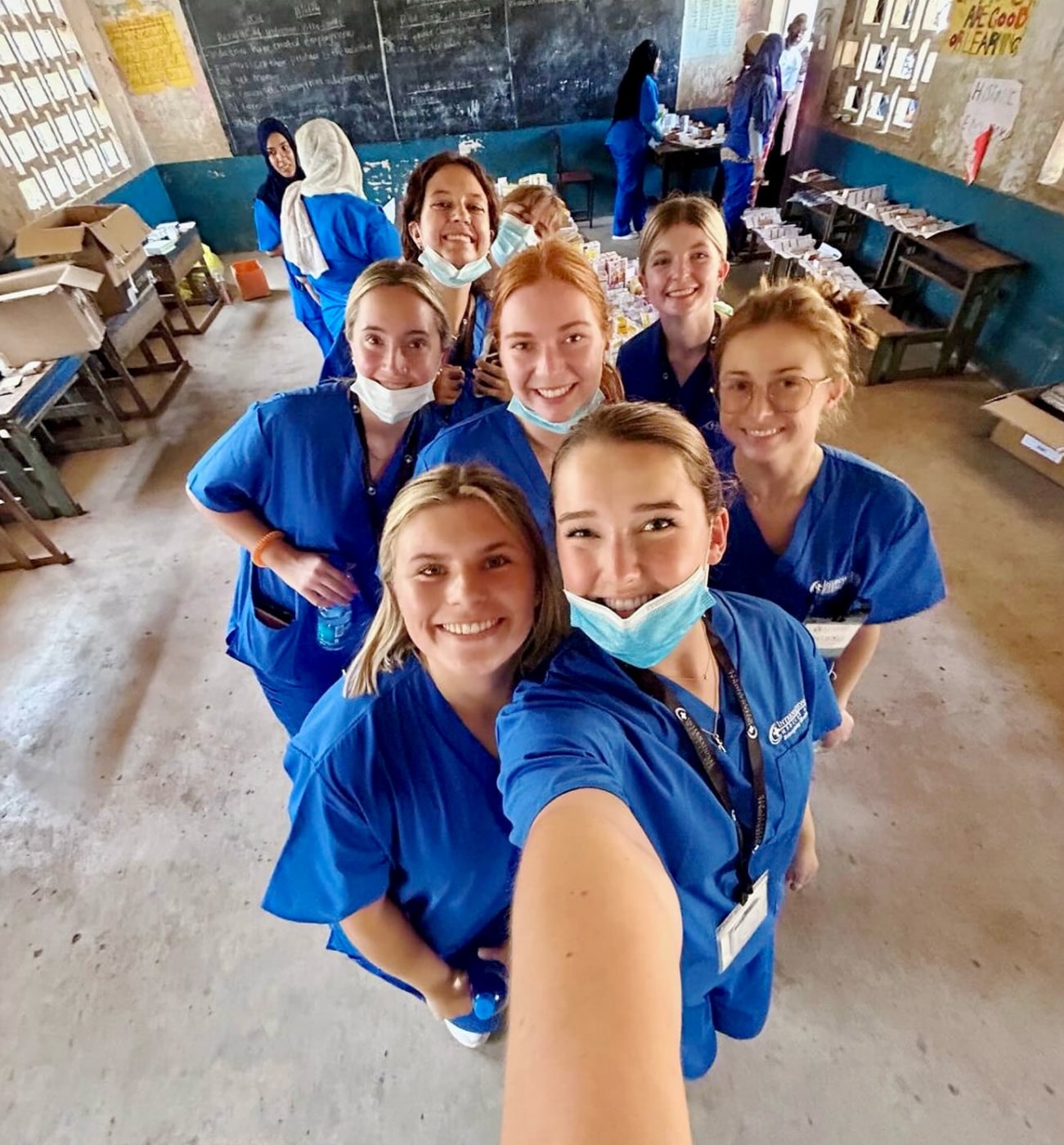 Group of cheerful medical interns in blue scrubs taking a selfie inside a classroom-turned-clinic.