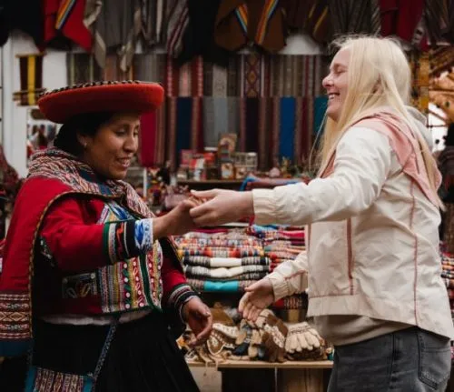 Smiling traveler engaging with a local artisan in traditional clothing at a colorful market stall showcasing handmade textiles and crafts.