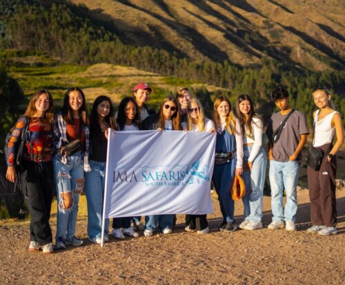 Group of young travelers posing outdoors with a “IMA Safaris” banner against a scenic mountain backdrop during an excursion.