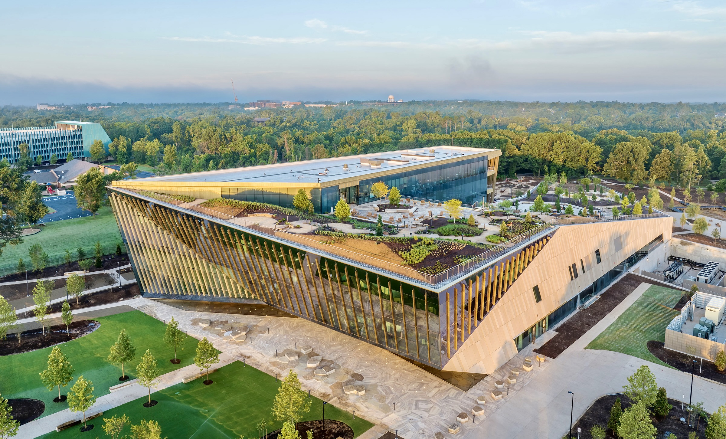 A modern, angular building with a green roof, surrounded by trees and pathways, likely part of a campus or complex.