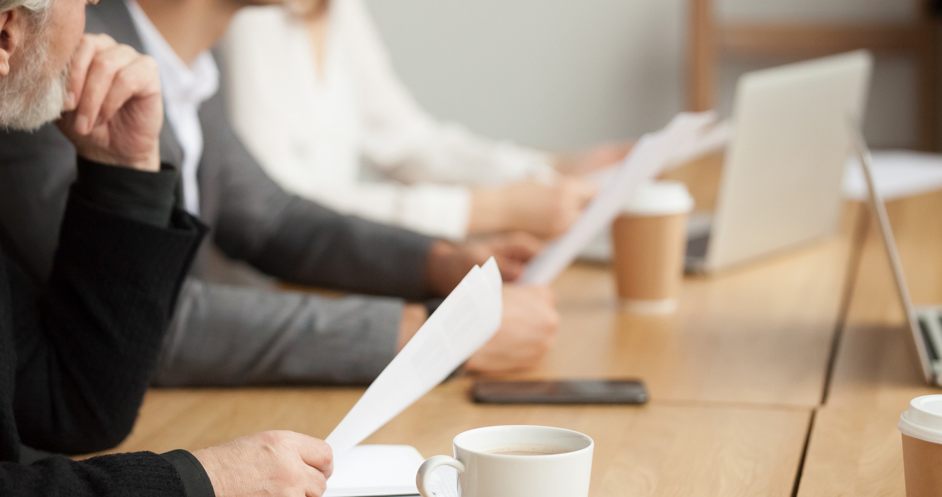 People are sitting at a meeting table with laptops, papers, and coffee cups.