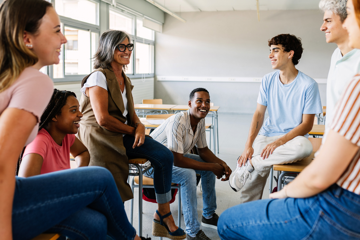 A group of people sitting and talking in a classroom setting, appearing relaxed and engaged.