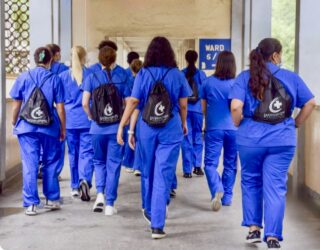 Group of medical interns in blue scrubs walking down a hospital corridor, wearing matching drawstring backpacks with the International Medical Aid logo.