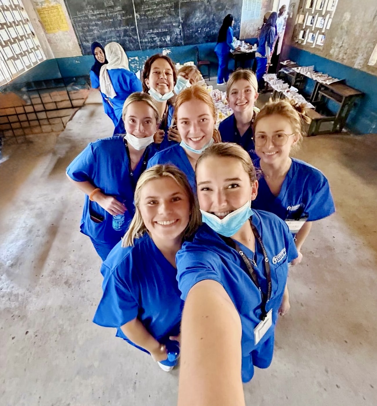 Group of cheerful medical interns in blue scrubs taking a selfie inside a classroom-turned-clinic.
