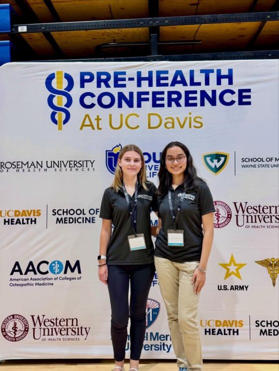 Two IMA representatives in black IMA polos standing in front of the Pre-Health Conference at UC Davis sponsor backdrop